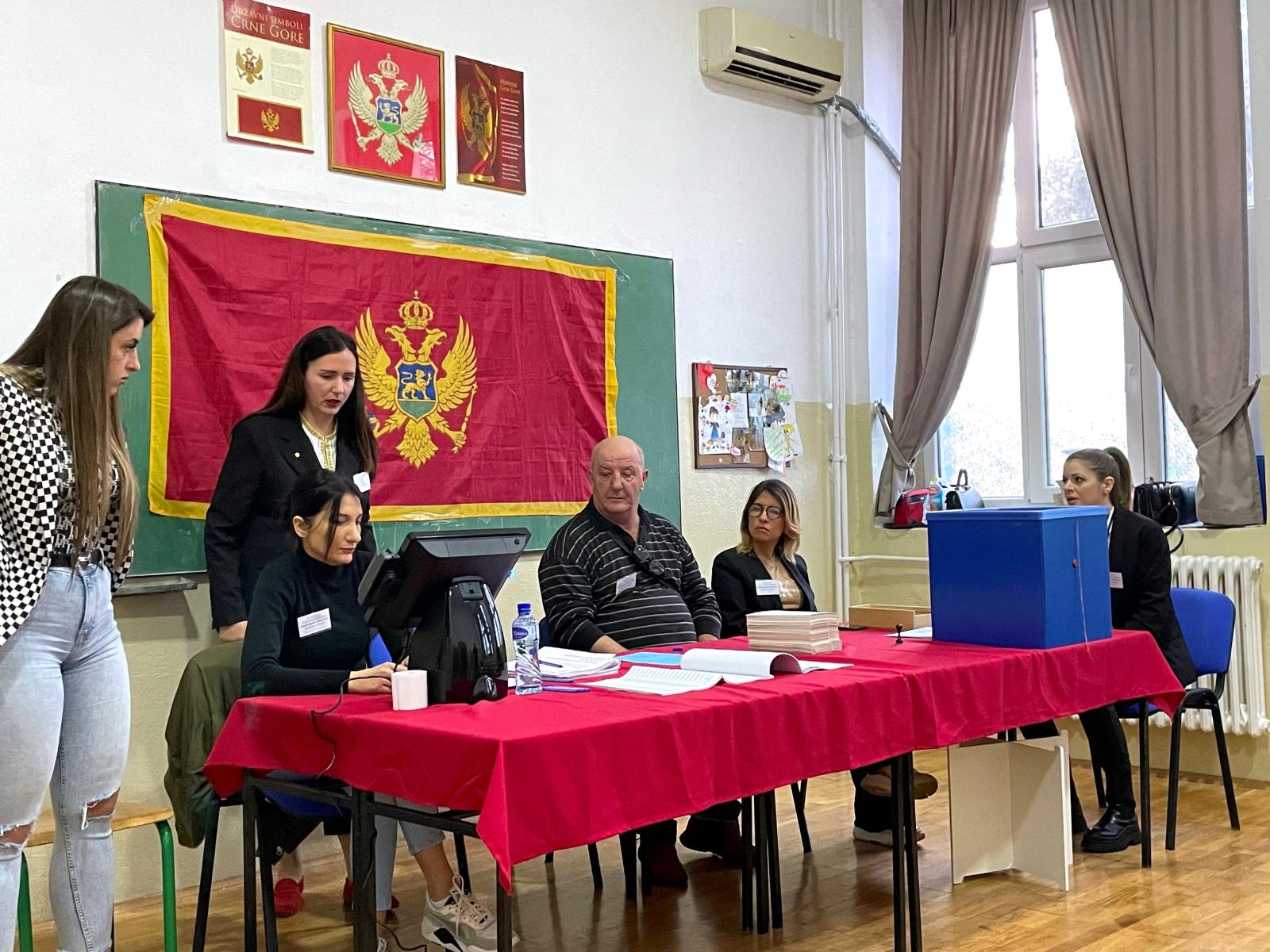 Four people sit at a red table, while two people stand behind the table. A Montenegro flag is hanging up on the wall behind them.